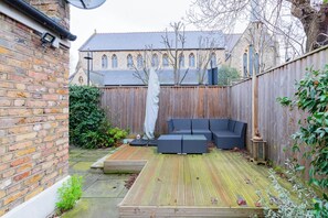 A wooden deck with a seating area, a brick wall, and a building with arched windows in the background. - Homely 2BD Flat with Private Garden, North London (Greater London)