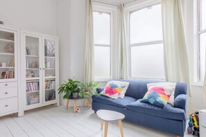 A room with a blue sofa, a white cabinet with glass doors, and a window with curtains. - Homely 2BD Flat with Private Garden, North London (Greater London)