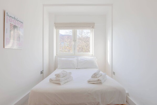 A hotel room with a bed, two pillows, a window with a view, and a poster on the wall.