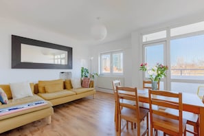 A modern living room with a sofa, dining table, chairs, and large windows. - Fantastically Located 4BD Flat, Clerkenwell (Greater London)