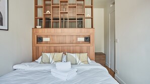 A neatly made bed with white linens and a folded towel, a wooden headboard with built-in shelves, and a small bedside table. - New Apartment Center of Paris/le Marais (Paris)