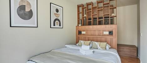 A neatly made bed with a white comforter and pillows, a wooden headboard with shelves, and two framed abstract artworks on the wall.