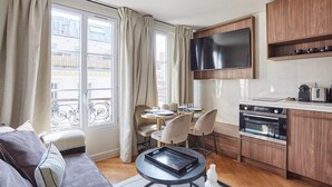 A modern living room with a sofa, a dining table with chairs, a flat-screen TV mounted above the kitchen counter, and a large window with curtains. - New Apartment Center of Paris/le Marais (Paris)