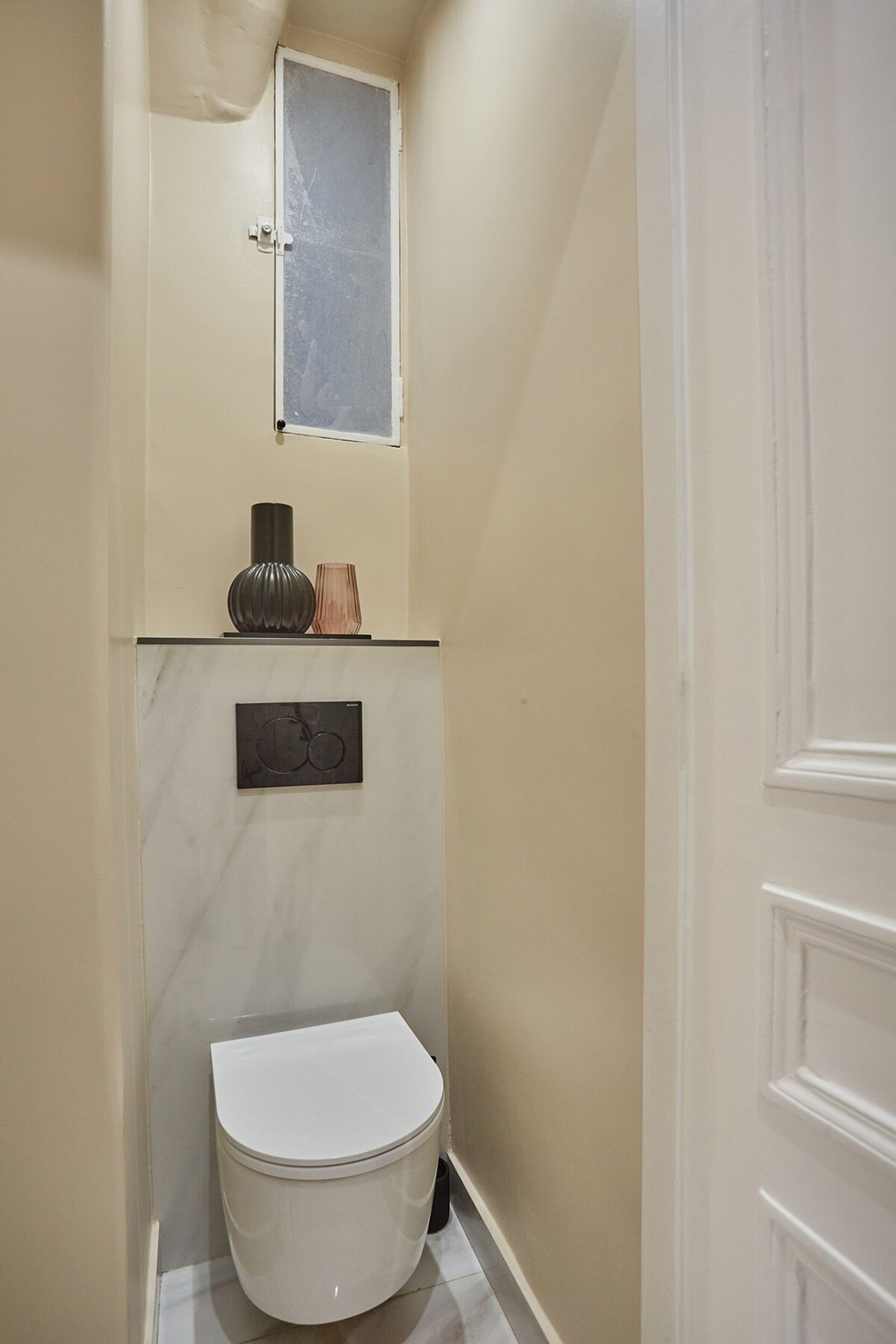 A narrow bathroom with a toilet, a small window, and a decorative shelf with a vase and a candle.