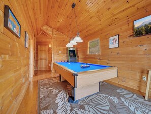 A wooden interior with a pool table, a ceiling light, and a view of the outdoors.