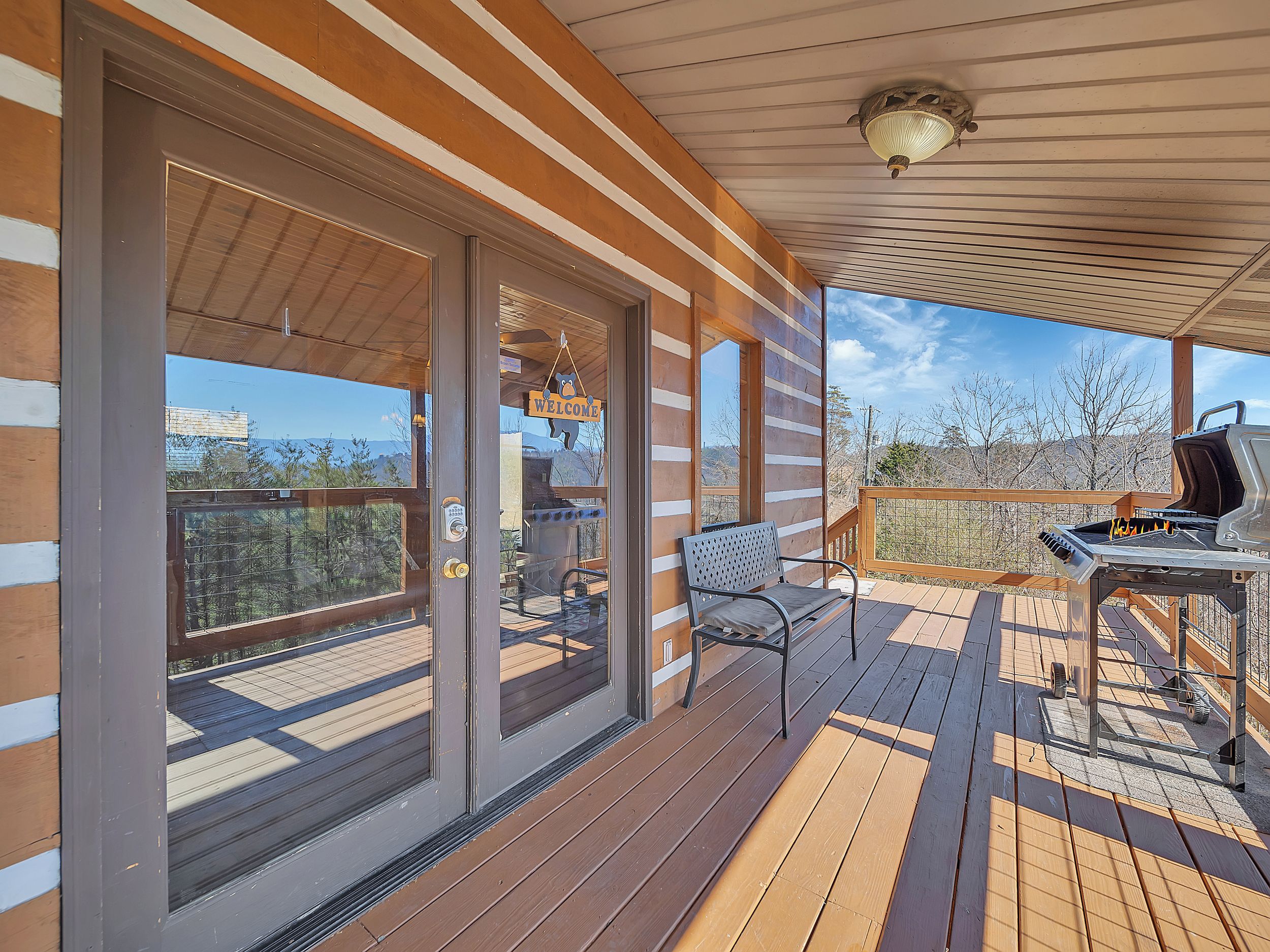 A wooden deck with a grill and a bench, a 'Welcome' sign, and a view of the mountains.