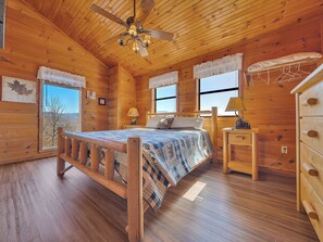 Un dormitorio tipo cabaña de madera con cama, mesita de noche, cómoda, ventilador de techo y ventana con vistas.