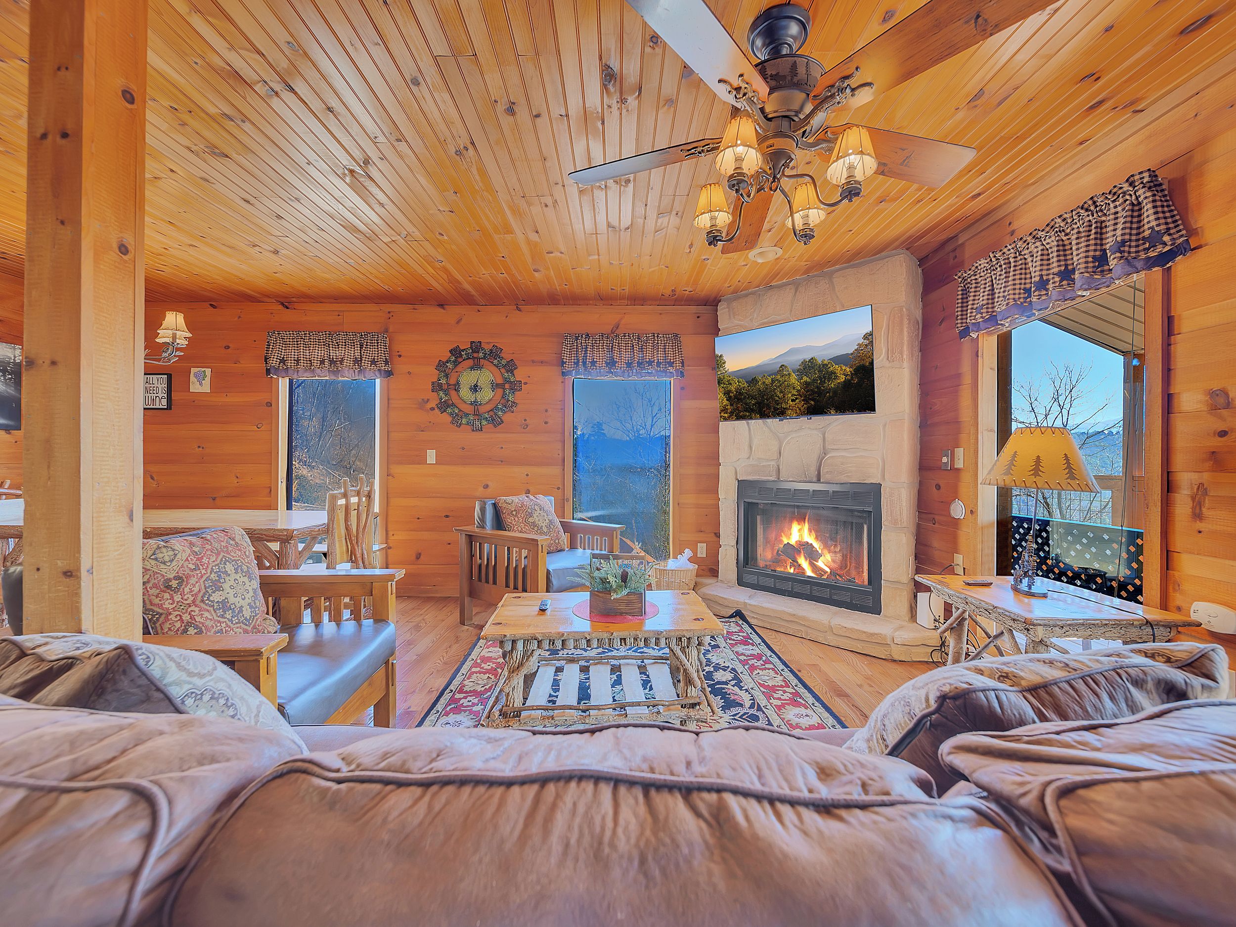 A cozy cabin interior with a fireplace, wooden walls, a ceiling fan, and a large window with a mountain view.