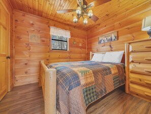 A wooden cabin bedroom with a bed, a window with a view of trees, a ceiling fan, and a wooden dresser.