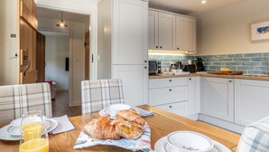 Eine moderne Küche mit weißen Schränken, einem Holztisch mit Frühstücksgebäck und Blick in den angrenzenden Raum.