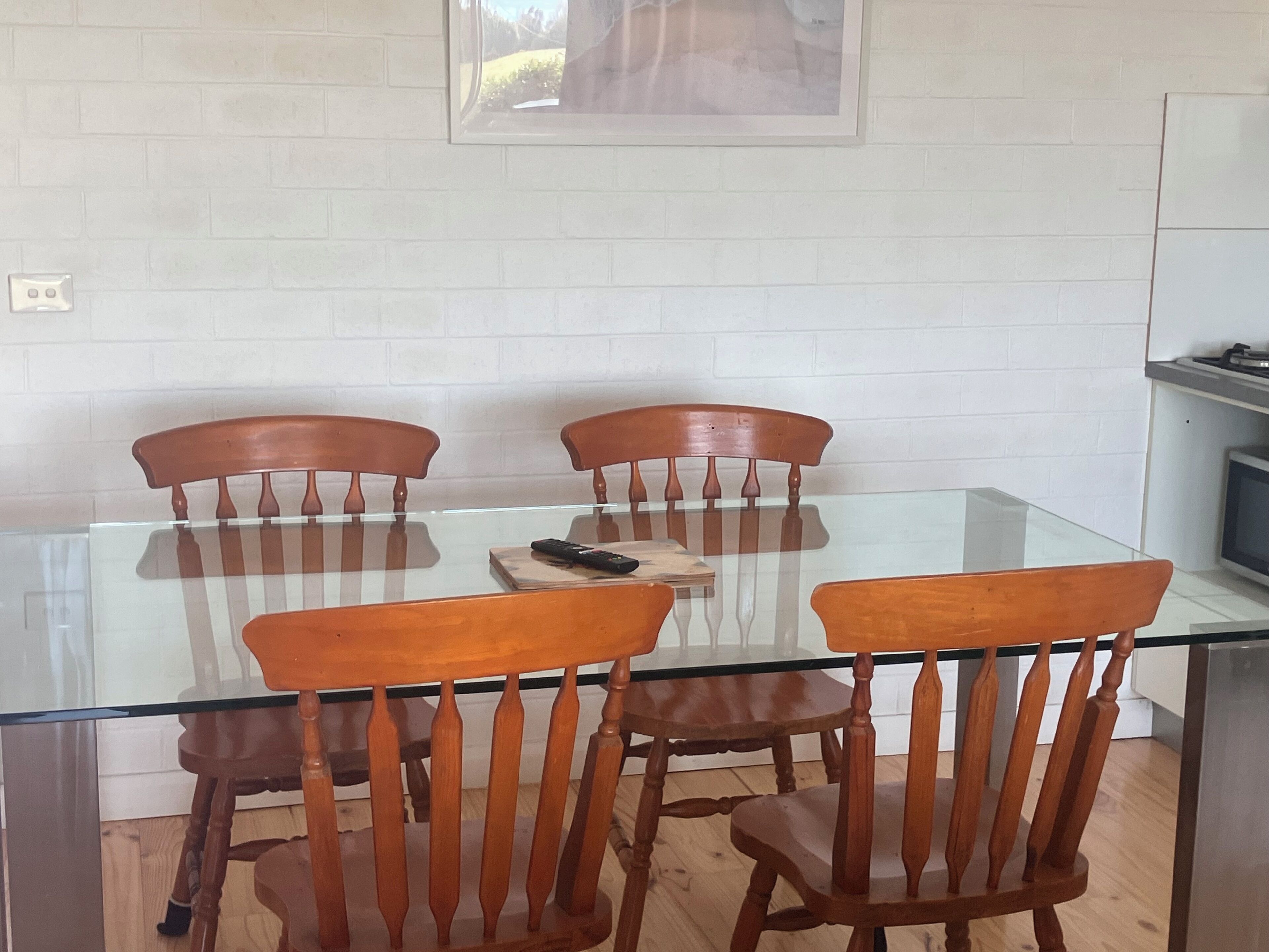 Een eetgedeelte met een glazen tafel en houten stoelen, een afstandsbediening op de tafel, en een witte bakstenen muur op de achtergrond.