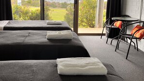 A hotel room with two beds, a small table, and a view of greenery and a road. - 61 OCEAN ROAD MOTEL  (Skenes Creek)