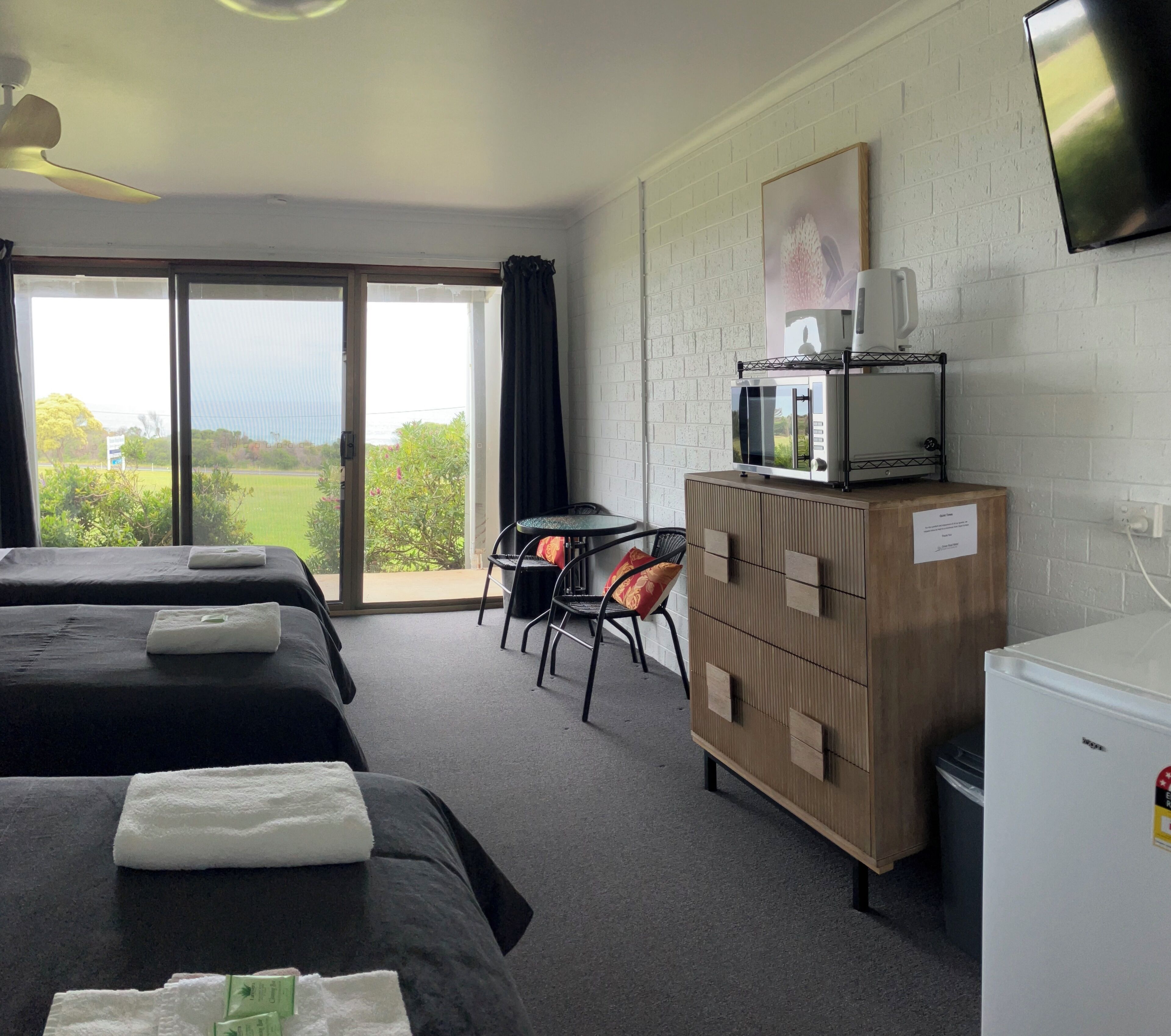 A hotel room with two beds, a desk, a chair, a microwave, a refrigerator, and a large window with a view of greenery.