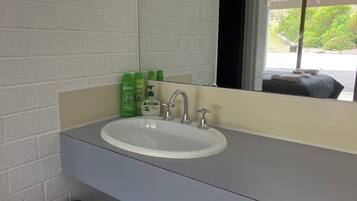 A bathroom with a white sink, green toiletries, and a large mirror reflecting a bed and greenery outside.