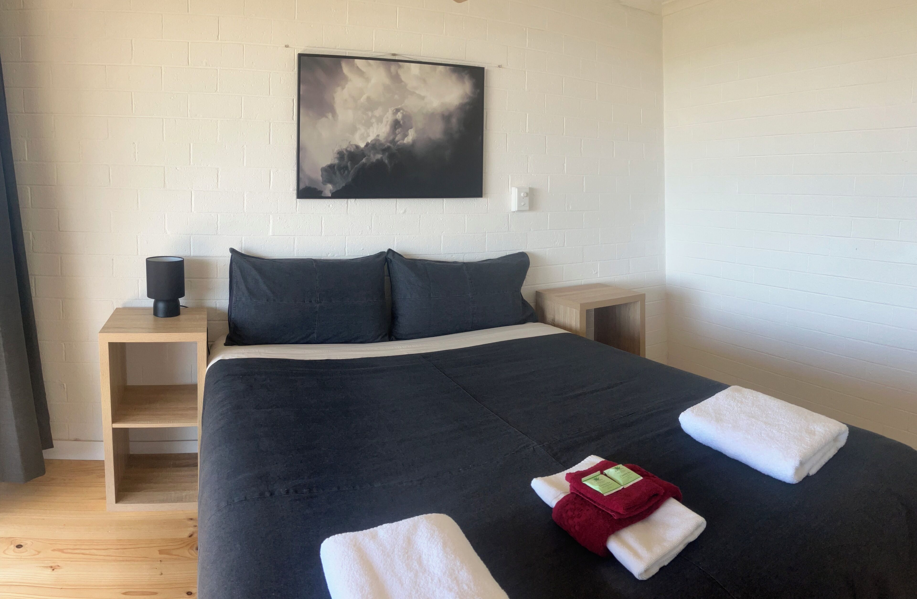 A neatly made bed with dark bedding, a side table with a lamp, and a painting on the wall.
