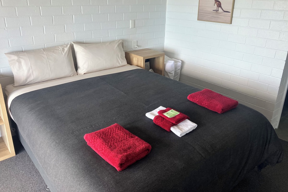Een netjes opgemaakt bed met een grijze deken, twee rode handdoeken en een witte handdoek op een donkergrijze vloer.