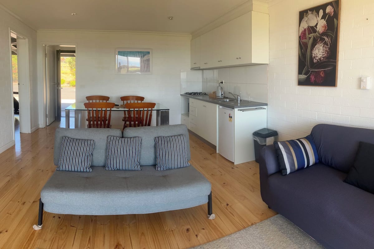 Een moderne woonkamer met een grijze bank, een donkere bank en een eethoek met houten stoelen en een glazen tafel.
