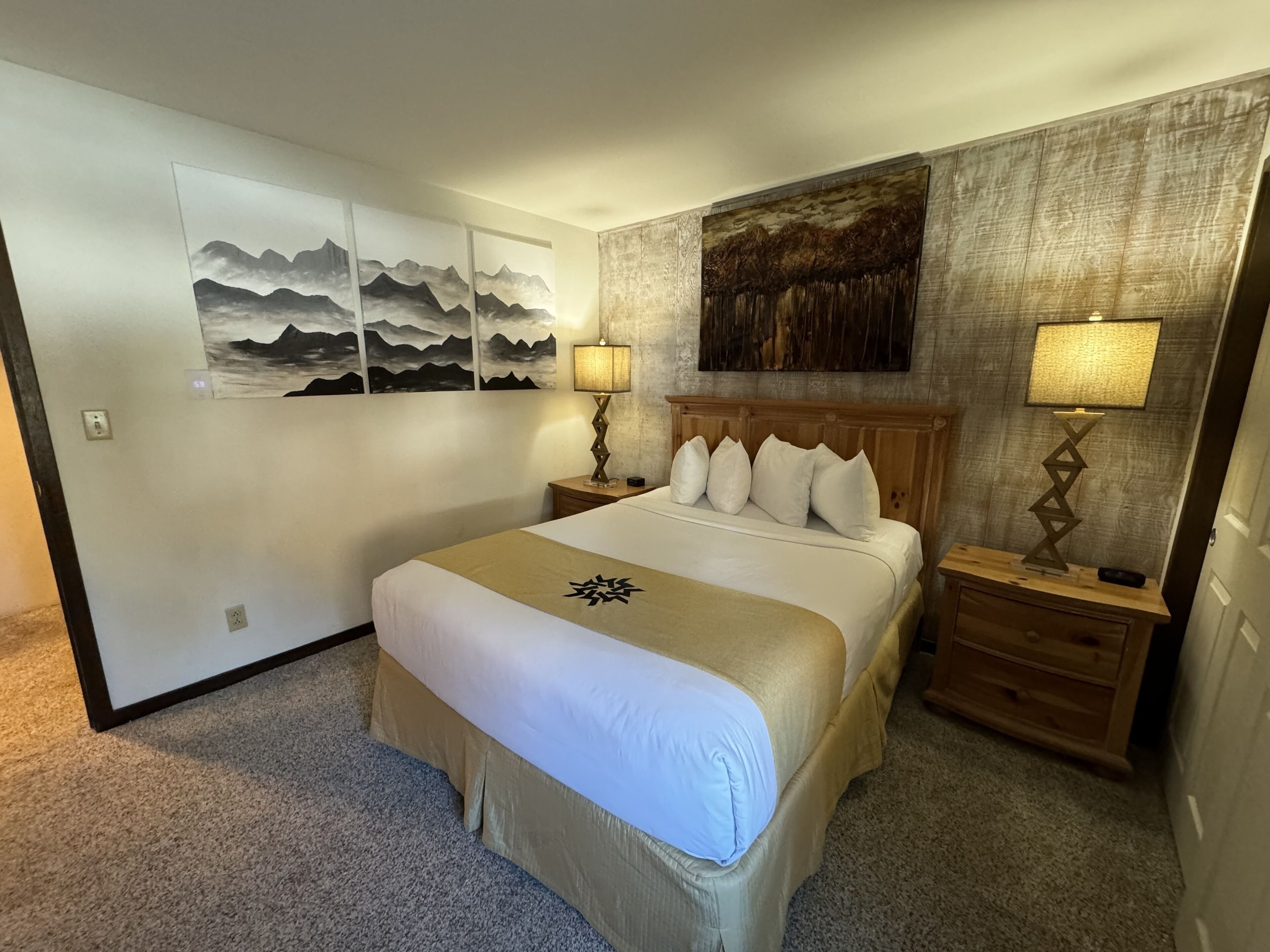 Une chambre d’hôtel avec un lit, deux tables de chevet, une lampe fixée au mur et un grand tableau au-dessus du lit.