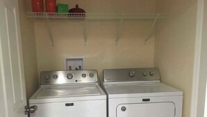 Un lavadero con dos lavadoras y secadoras apiladas, un estante arriba con productos de limpieza y un enchufe en la pared.
