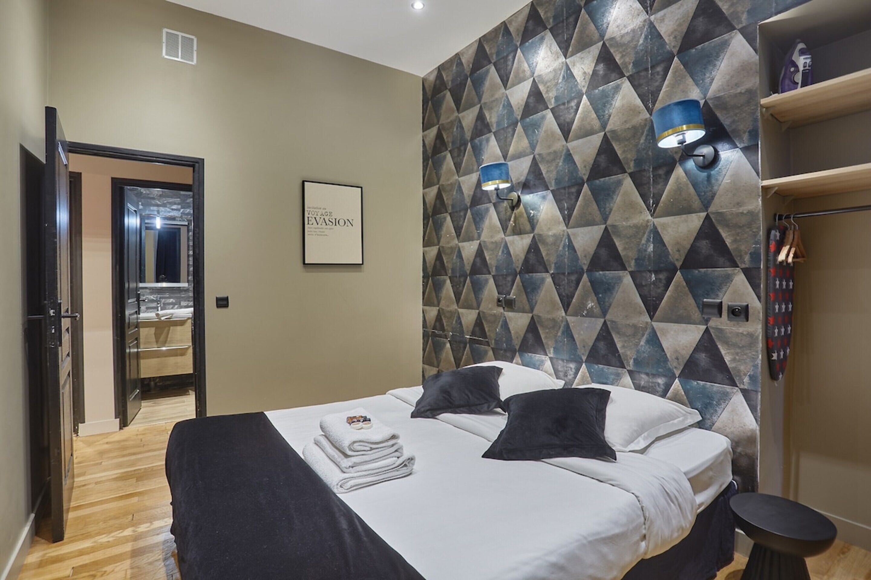 A hotel room with a bed, a wall with geometric patterns, a door leading to a bathroom, and a shelf with toiletries.