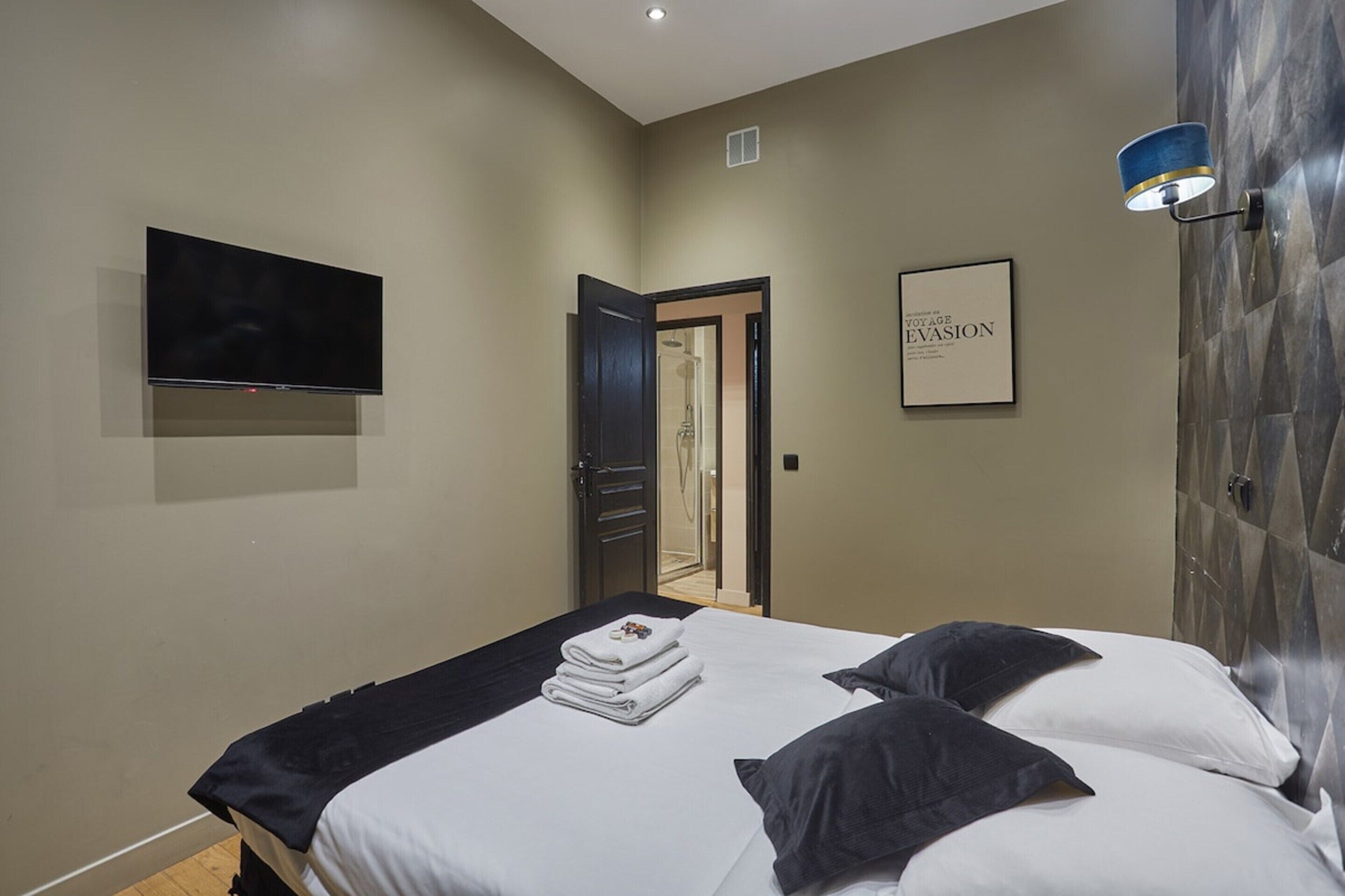 A bedroom with a bed, a flat-screen TV mounted on the wall, a framed picture, and a door leading to another room.