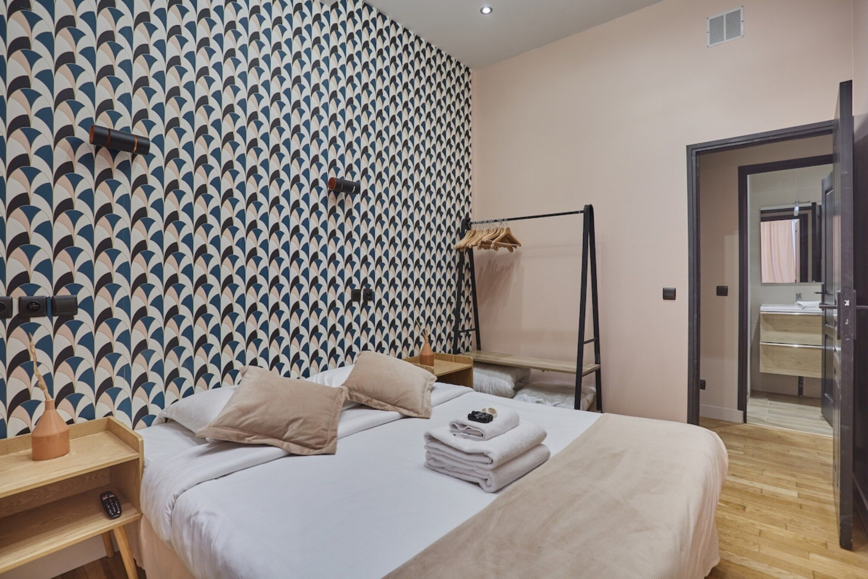 A neatly arranged hotel room with a patterned wall, a bed with pillows and towels, a wooden nightstand, and a wardrobe.