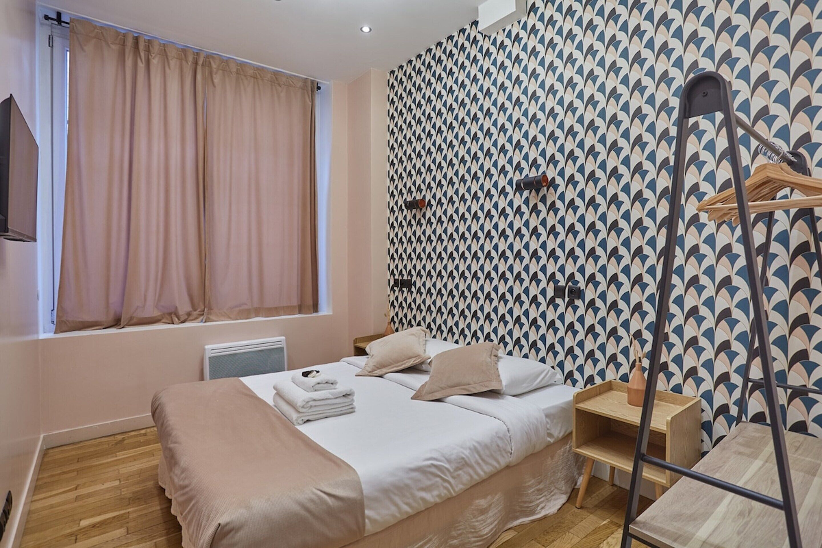 A hotel room with a bed, a window with curtains, a radiator, a wooden floor, a patterned wall, and a wooden hanger stand.