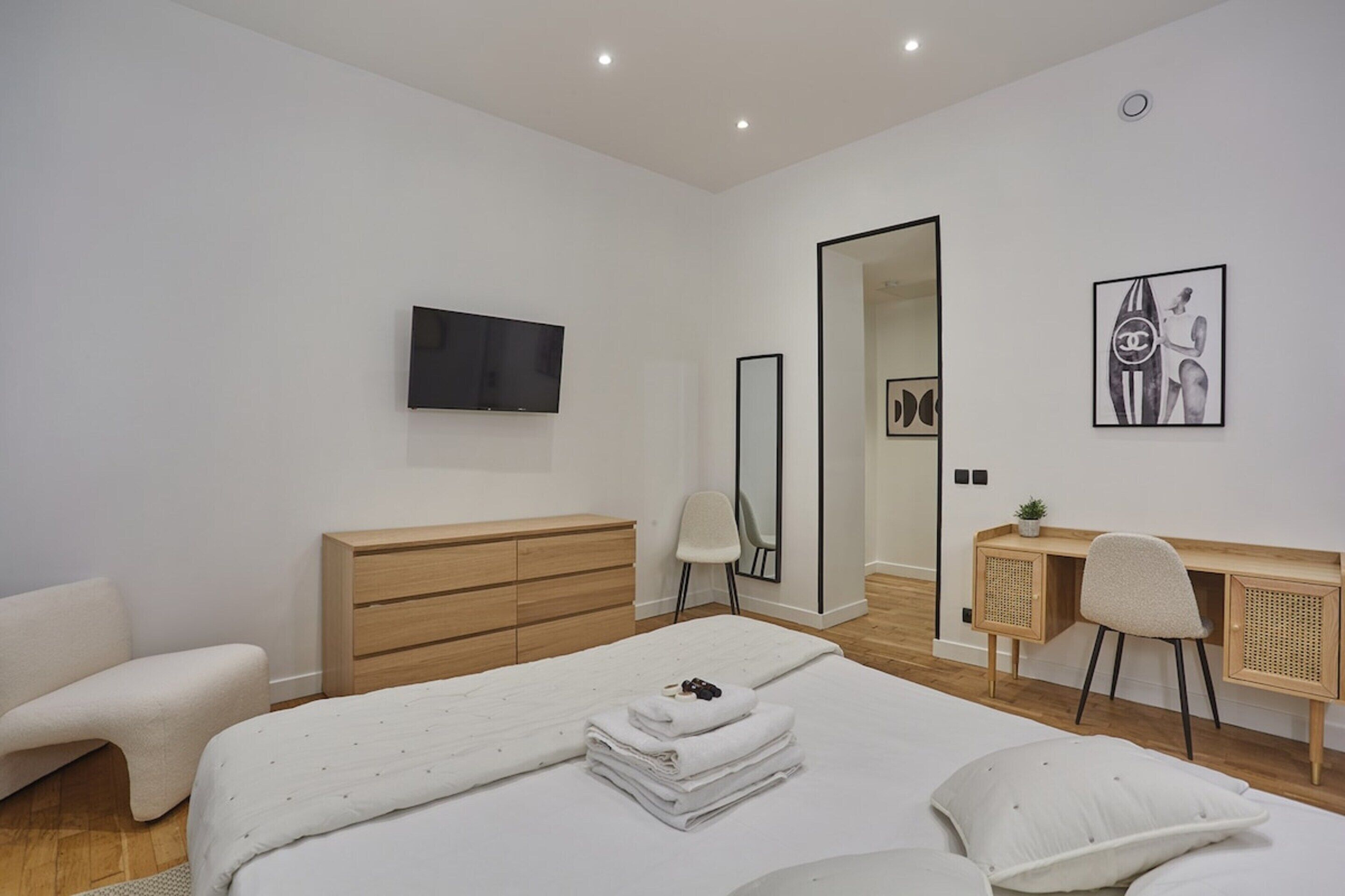 A modern bedroom with a bed, a wooden dresser, a desk with a chair, a TV mounted on the wall, and a large mirror.