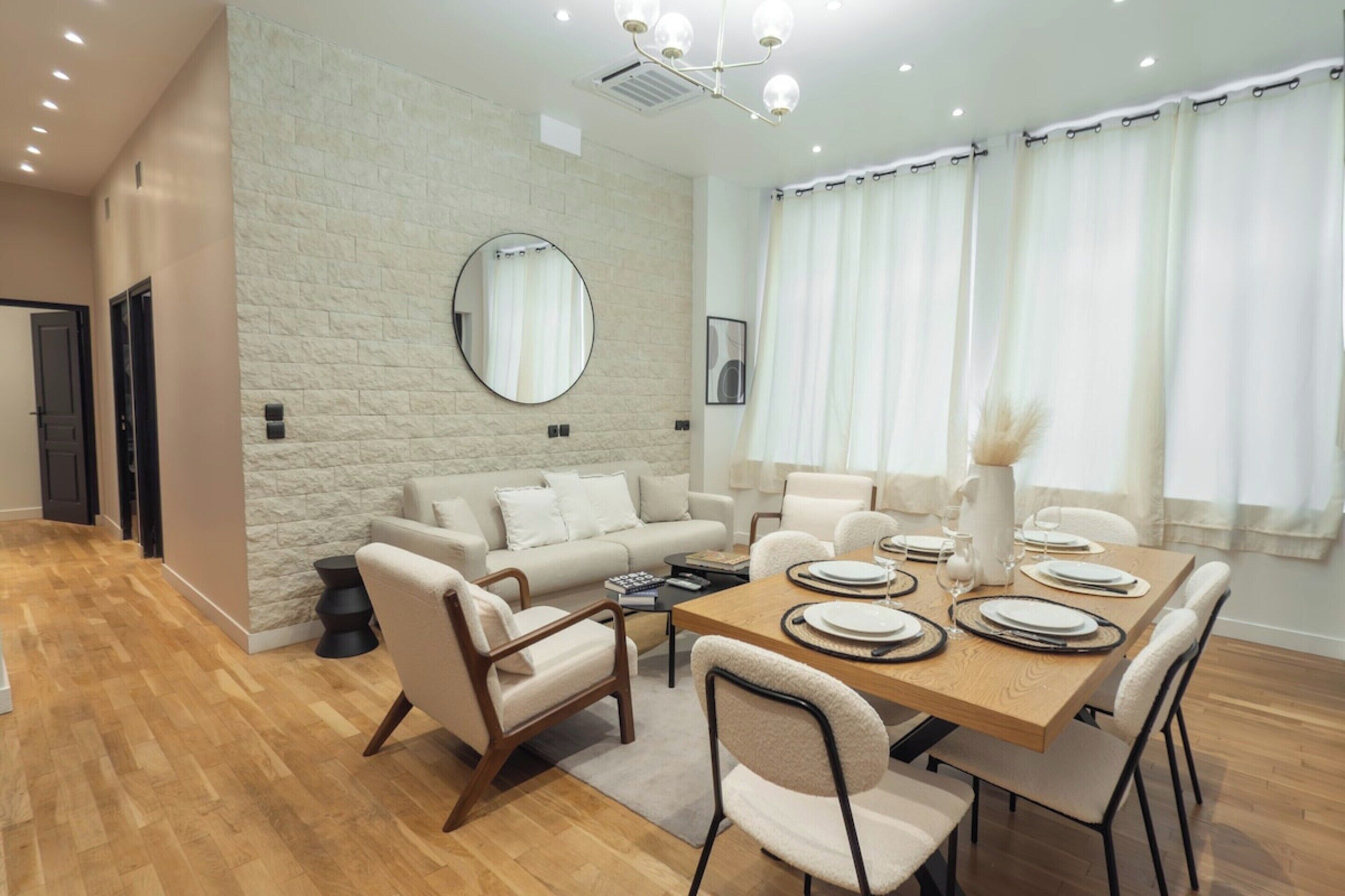Uma sala de jantar moderna com uma mesa de madeira preparada para quatro pessoas, rodeada por cadeiras, num espaço com parede de pedra e um espelho grande.