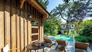 A wooden deck with lounge chairs and a table, overlooking a swimming pool and a house in the background. - Cap Ferret Villa • 5 Bedrooms • Heated Pool (Lège-Cap-Ferret)