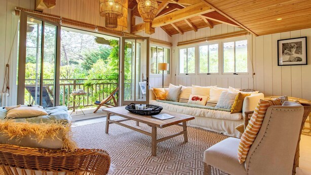 A cozy room with a wooden ceiling, large windows, a sofa, and a wicker chair. - Cap Ferret Villa • 5 Bedrooms • Heated Pool (Lège-Cap-Ferret)