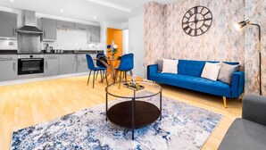 A modern kitchen with grey cabinets and a dining area with a round glass table and blue chairs. A blue sofa and a floor lamp are in the living room.