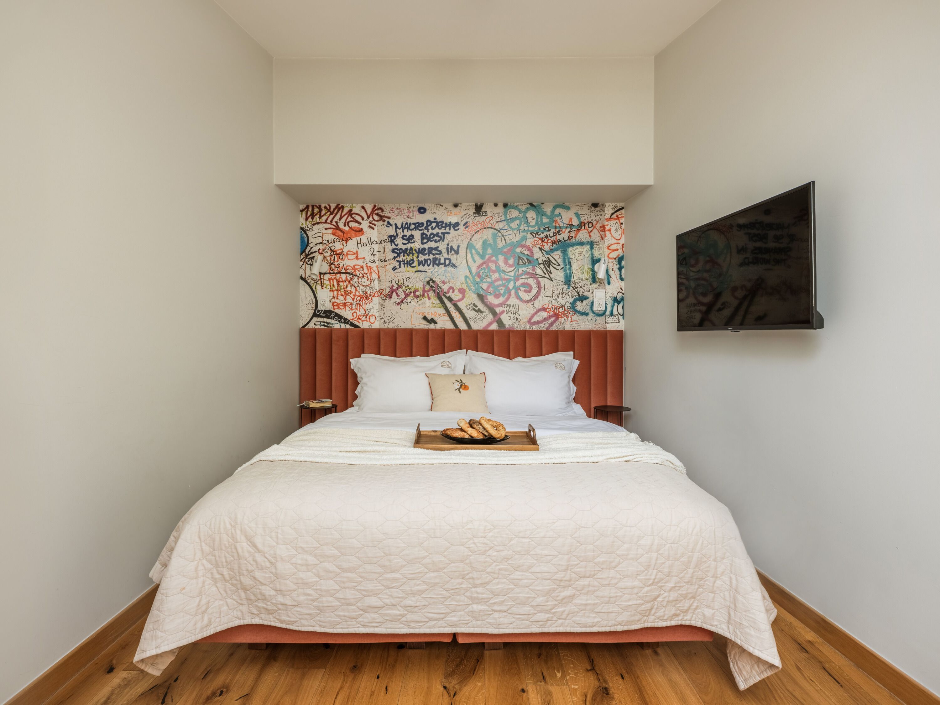 A bedroom with a bed, a wooden floor, a white quilted bedspread, a wooden headboard, and a graffiti-covered wall.