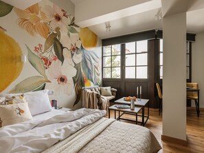 A bedroom with a large floral mural, a bed with white bedding, a bedside table with books, and a view of the outside through a window. - Studio standard for two. (Kraków - Kazimierz & Podgórze)