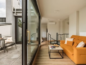 Un salon moderne avec une porte vitrée donnant sur un balcon, un canapé, une table basse et une petite table sur laquelle sont posés des livres et des pâtisseries.