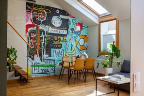 Ein Zimmer mit geneigter Decke, einem Wandgemälde mit abstrakter Kunst, einem Essbereich mit Stühlen und Tisch sowie einem Fenster mit Blick.