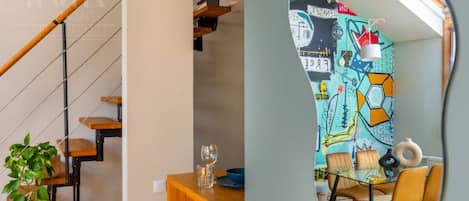 Una escalera con escalones de madera y barandillas de metal, un comedor con mesa y sillas, y una pared colorida con diseños abstractos.
