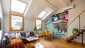A loft-style living room with a high ceiling, skylights, a colorful mural, a staircase, and a dining area.