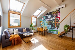 A loft-style living room with a high ceiling, skylights, a colorful mural, a staircase, and a dining area.