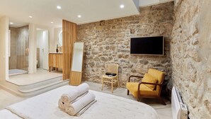 Una moderna camera d'albergo con una parete in pietra, un letto con asciugamani arrotolati, una sedia in legno, una televisione e un bagno con doccia e WC.