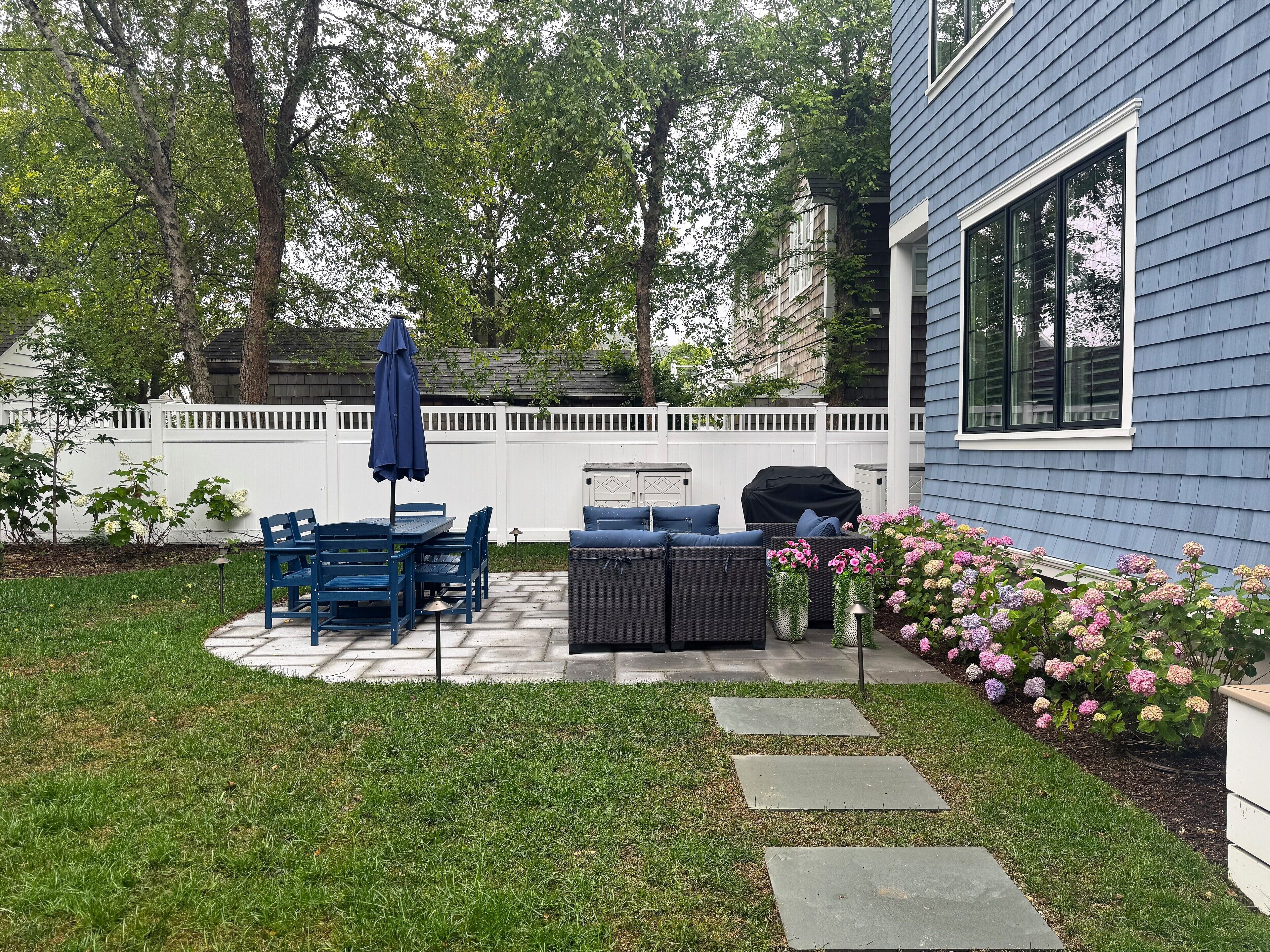 A backyard with a patio, outdoor furniture, a grill, and a white fence.