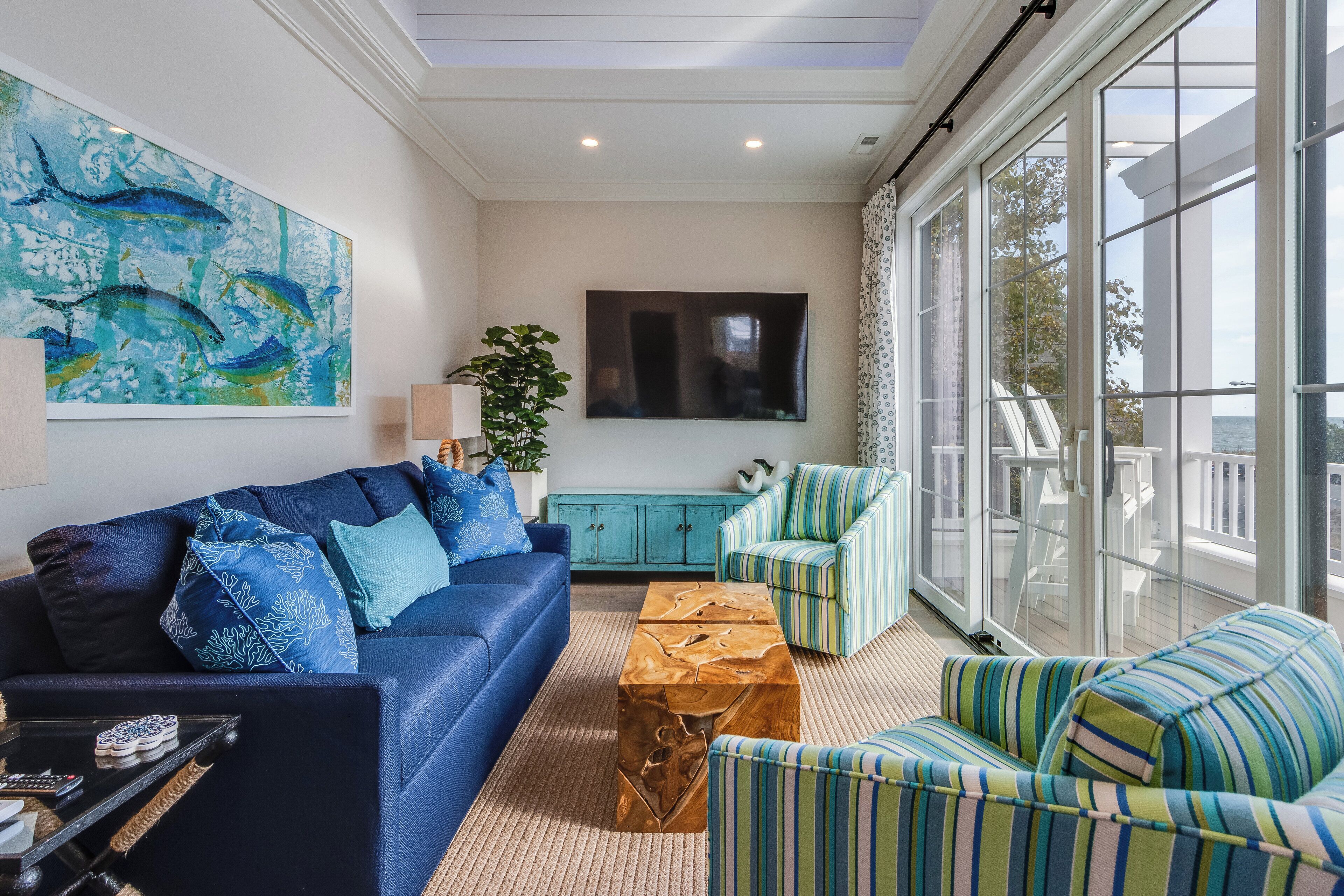 A modern living room with a blue sofa, striped armchairs, a wooden coffee table, and a large window offering a view of the outdoors.