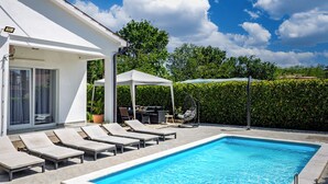 En swimmingpool med solstolar, en möblerad sittgrupp under tak och en vit byggnad med en glasdörr.