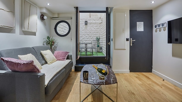 A modern living room with a grey sofa, a small table with a bottle and glasses, and a view of an outdoor patio through an open door. - Charming Apartment Faubourg St Honore Madeleine (Paris)