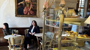 A cafe interior with a woman seated at a table, a glass display case with pastries, and a classical painting on the wall.