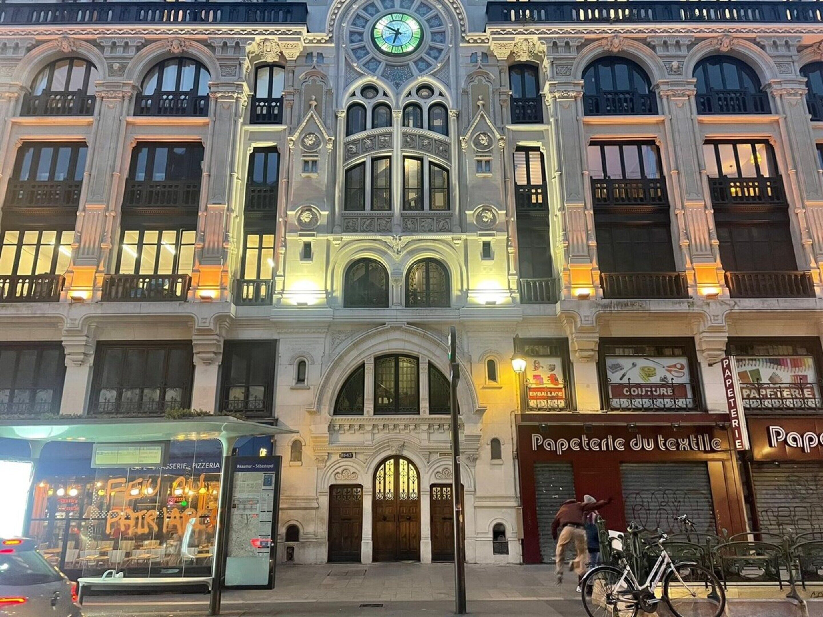 Un bâtiment historique à l’architecture riche et élaborée, qui se distingue par une horloge imposante et abrite plusieurs boutiques, dont une boulangerie et une boutique de vêtements.