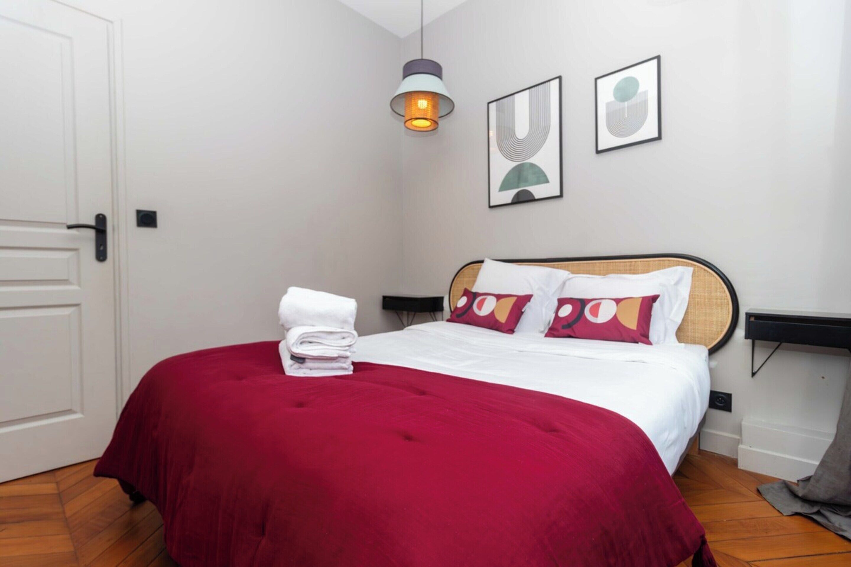 A neatly made bed with a red bedspread, white pillows, and a folded towel on the side. There is a wall-mounted light fixture above the bed and two framed artworks on the wall.