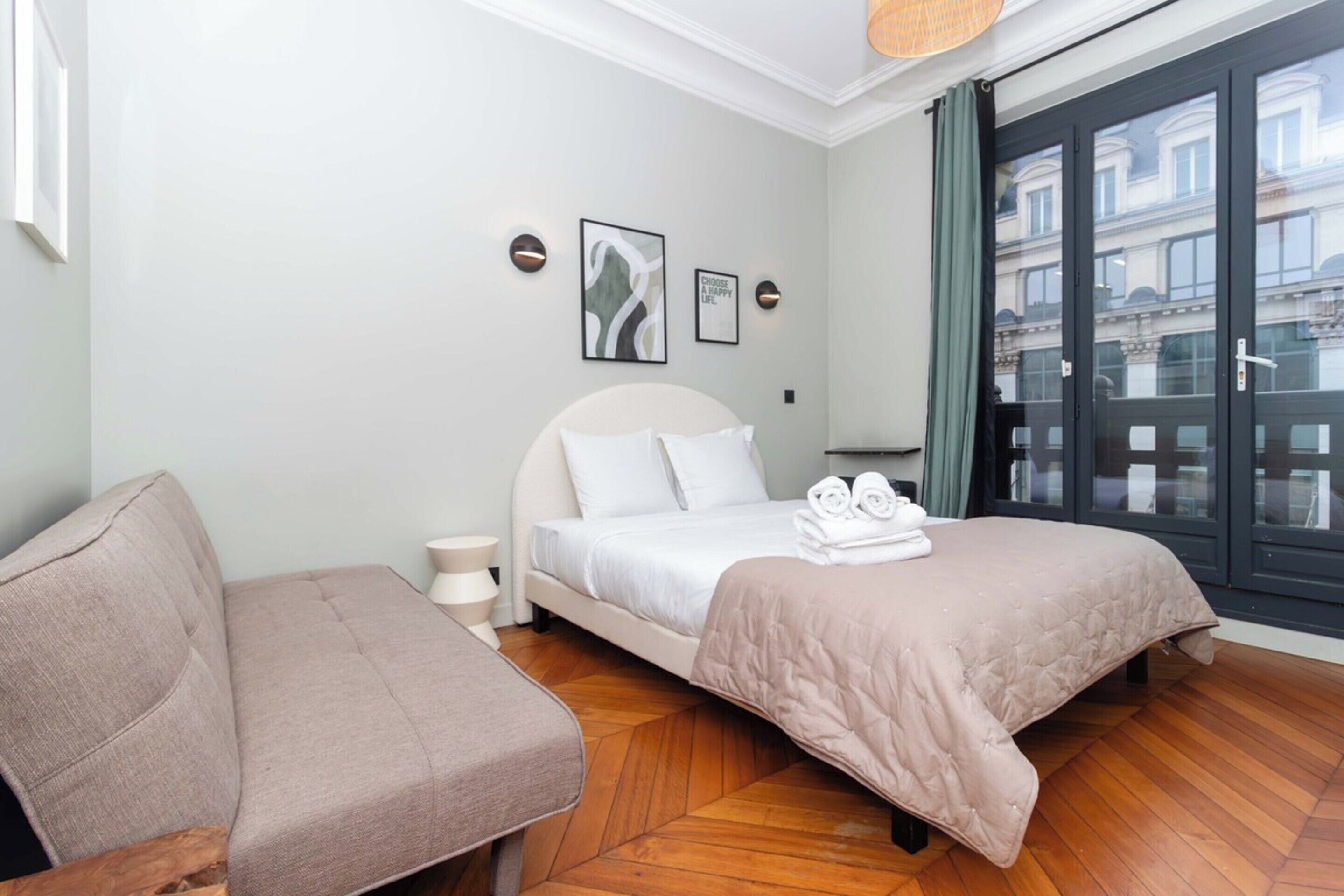 Ein Schlafzimmer mit Bett, einer Couch und Blick auf Gebäude durch ein großes Fenster.