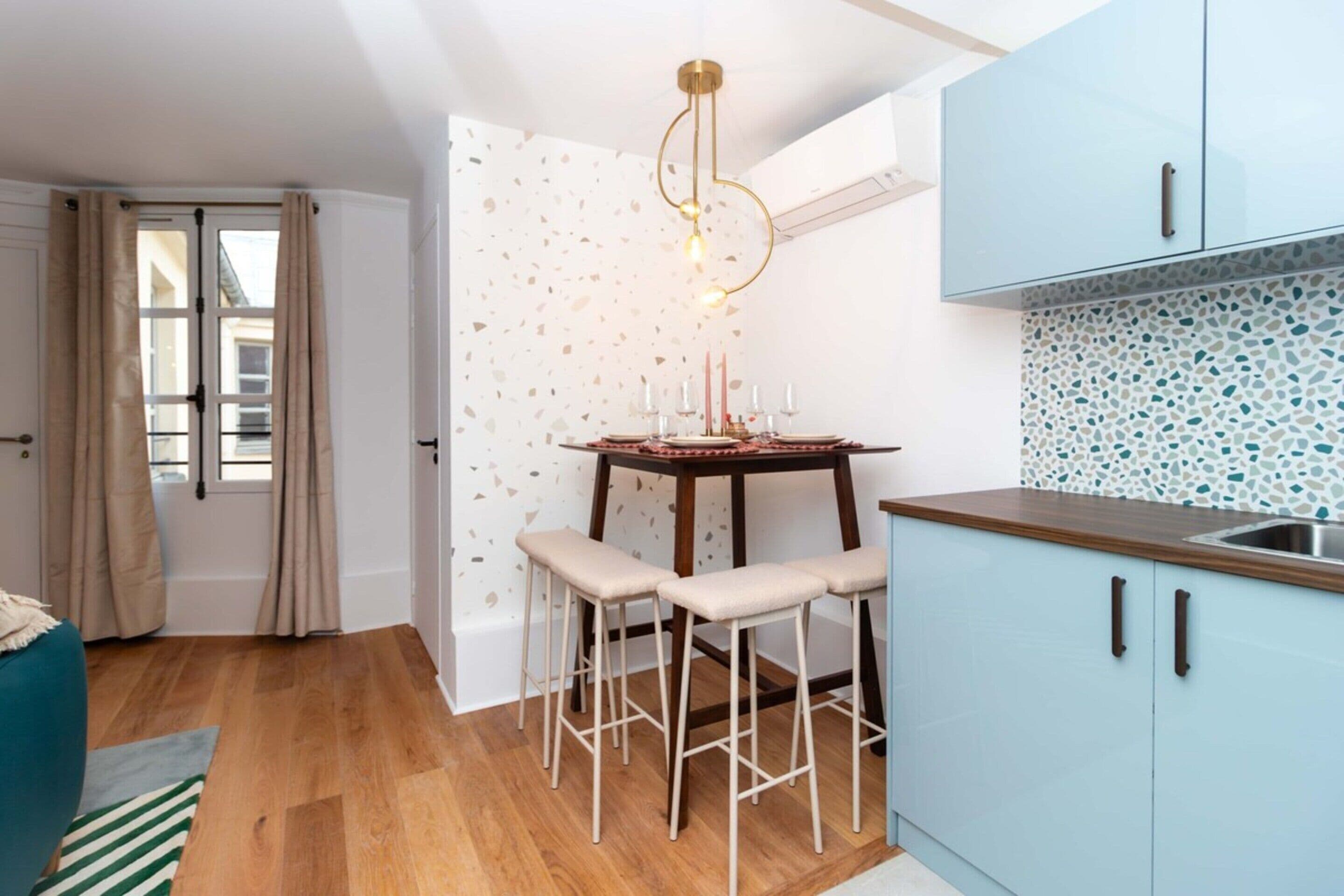 A compact kitchen with a dining area, featuring a small table with stools, a hanging light fixture, and light blue cabinets.