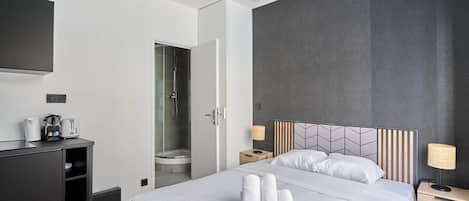 Una habitación de hotel moderna con una cama grande, zona de ducha, televisor de pantalla plana, escritorio y mesita de noche con lámparas.